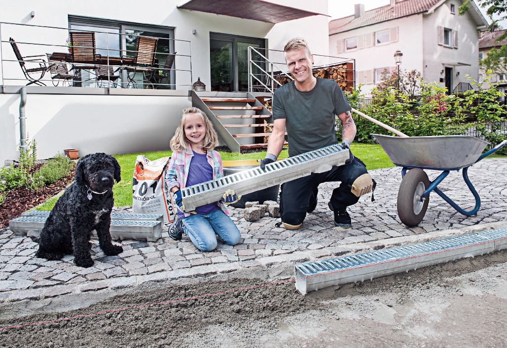 Vater baut mit seiner kleinen Tochter auf der Terrasse eine Terrassenentwässerung ein. Es handelt sich um eine Entwässerungsrinne. Ein Hund sitzt dabei.