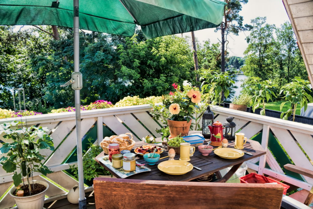 Ein Balkon mit vielen bunten Blumen. Ein Holztisch ist mit Geschirr und Lebensmitteln gedeckt (ökologisch bauen).