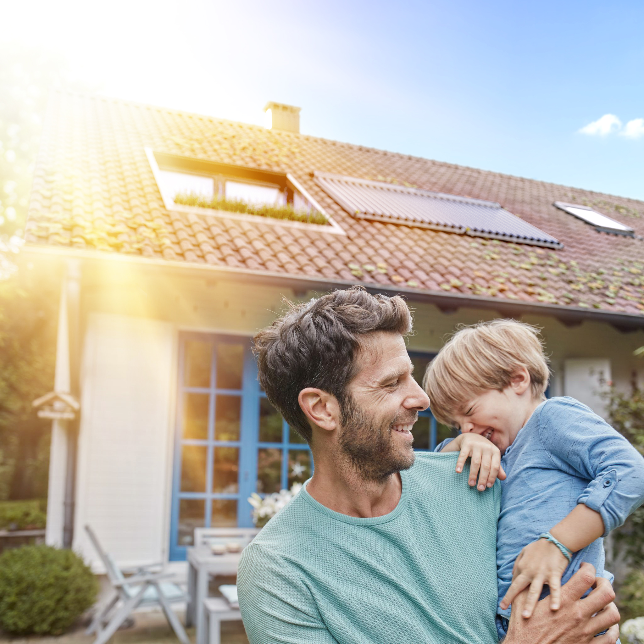 Glückliche Familie vor Haus mit Solartechnik Vater mit Kind vor Einfamilienhaus mit Solaranlage auf dem Dach bei Sonnenschein